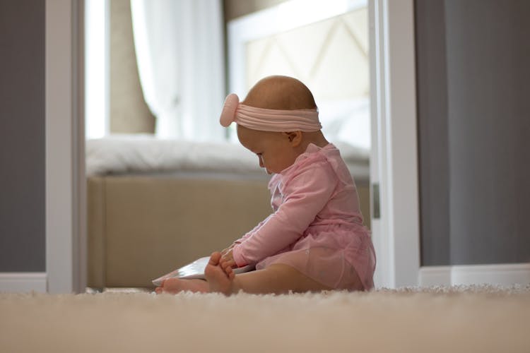 Baby In Pink Dress And Headband Sitting On A Rug