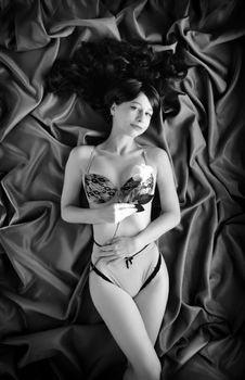 Top view of black and white alluring lady in sexy lingerie lying on silk bed sheet with rose in hand and looking at camera