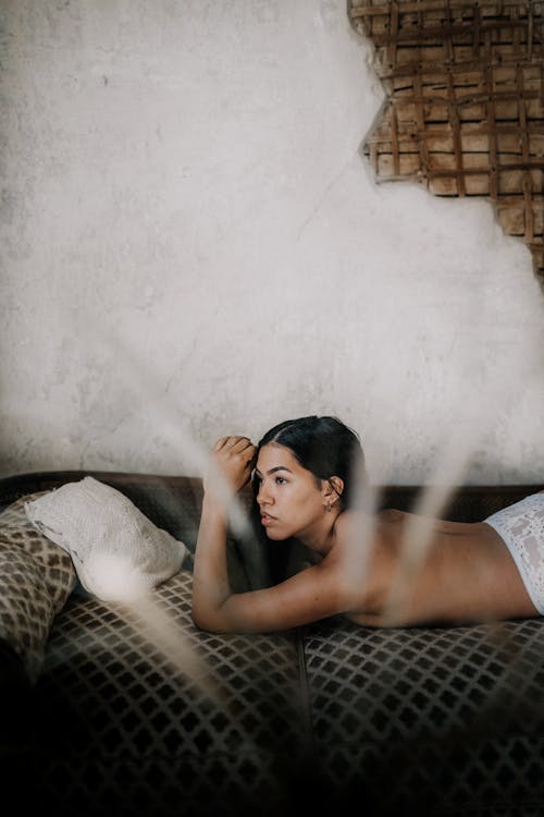 Free Side view of young dreamy topless ethnic female lying on couch with decorative cushions and looking away Stock Photo