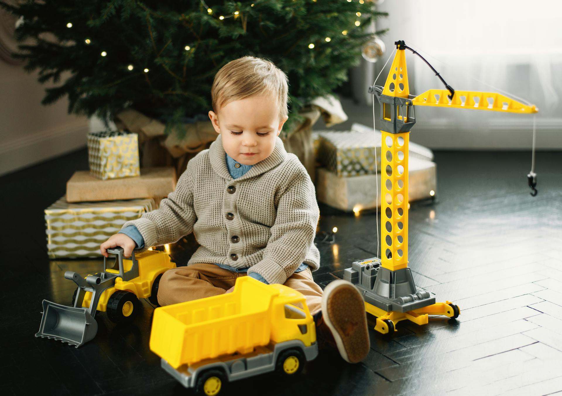 A young boy plays with toy trucks and a crane under the Christmas tree.