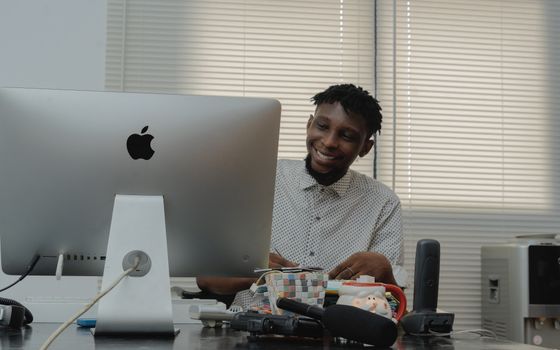 African man smiling while working at a desk with a computer in a well-lit office space.