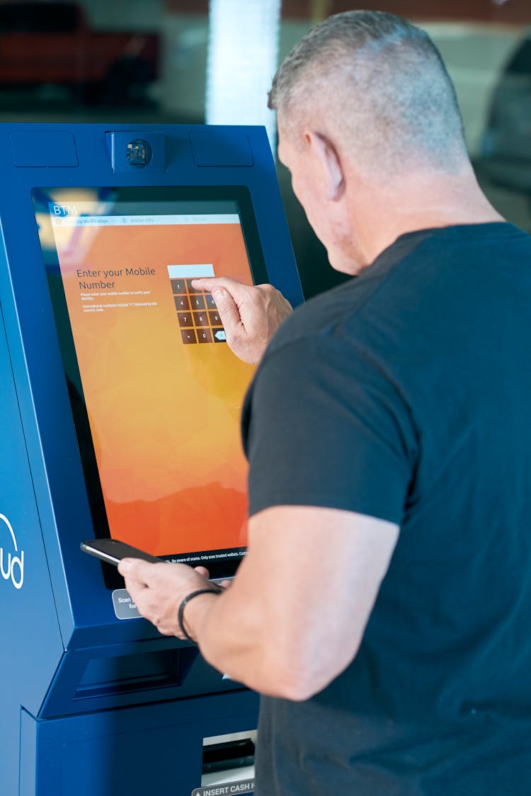 Man Holding A Mobile Phone While Touching An ATM Screen