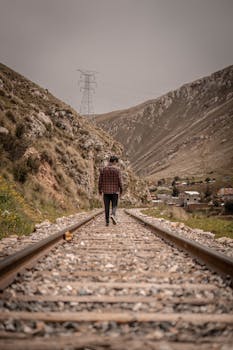 A man in a plaid shirt walks along rustic railroad tracks in the scenic mountainous terrain of Peru.