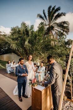 Young diverse couple in bridal apparel holding hands on carpet with anonymous black male marriage officiant and guests during festive event