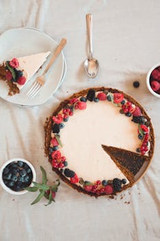 Top view of a berry tart with fresh fruits and cream, styled with plates and bowls on a white surface.