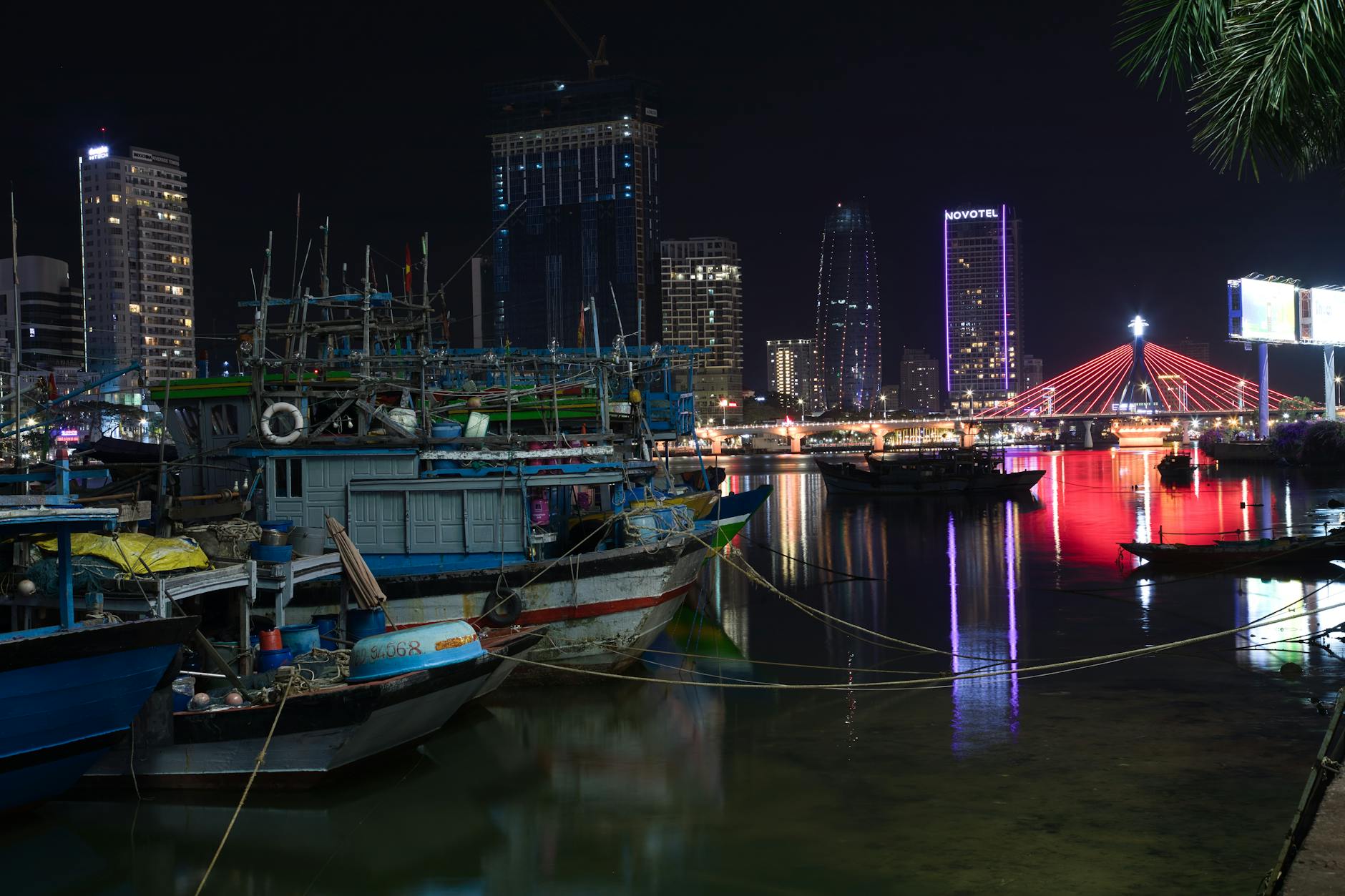 Illuminated cityscape of Đà Nẵng at night featuring boats and a vibrant bridge reflection.