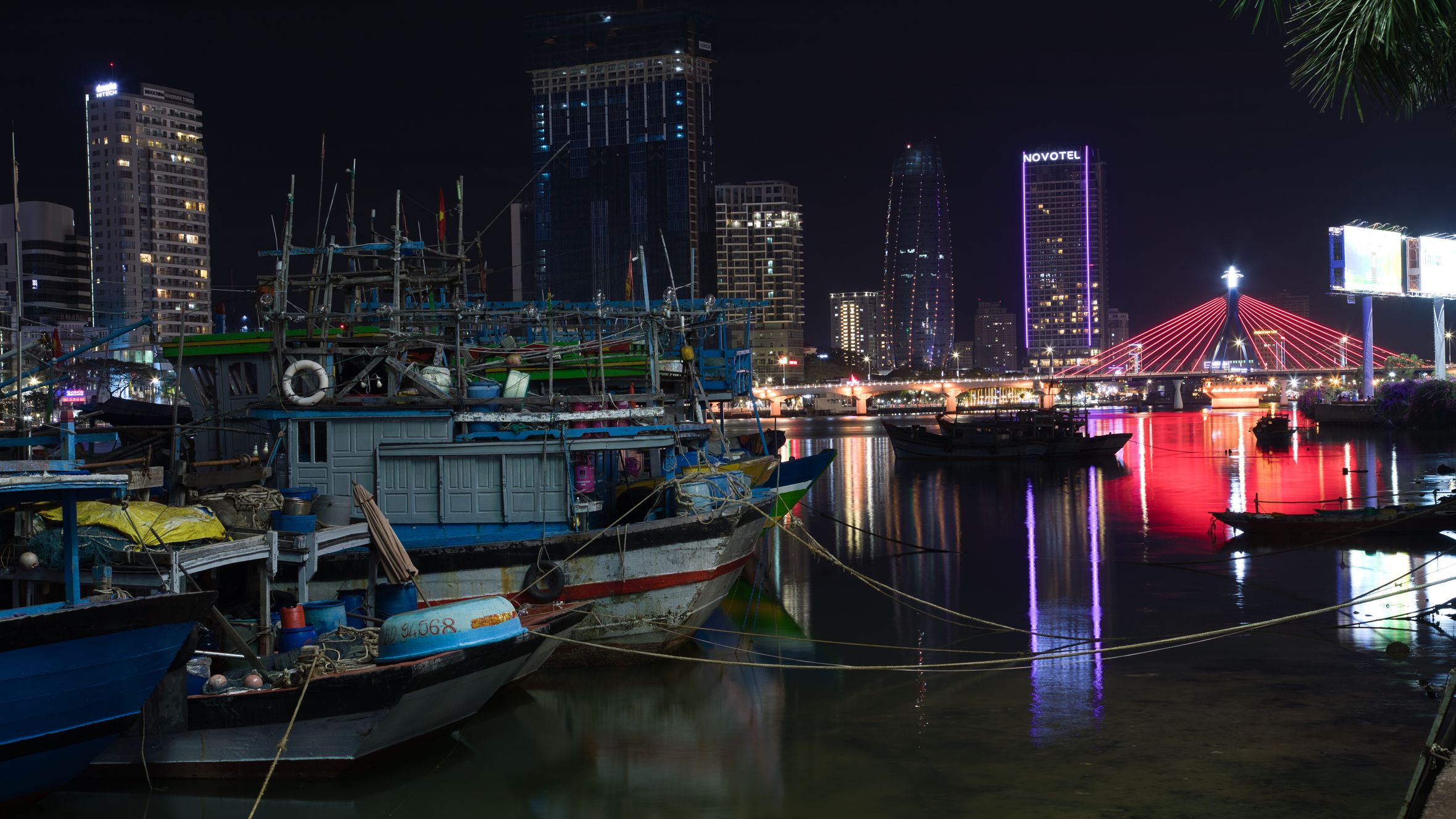 Illuminated cityscape of Đà Nẵng at night featuring boats and a vibrant bridge reflection.