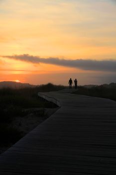 Two people walking on a boardwalk at sunset in Galicia, Spain with a stunning sky backdrop.