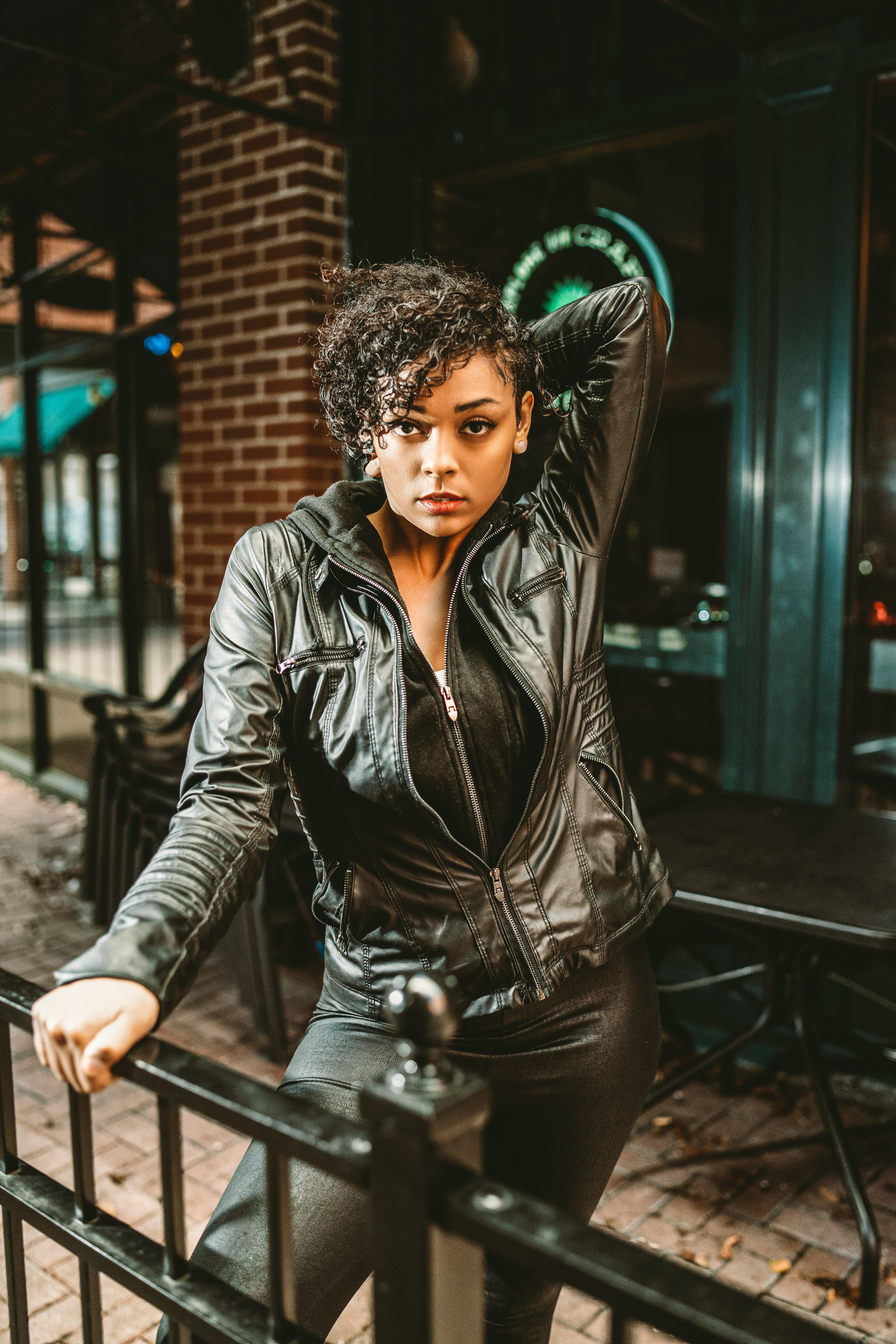 Young informal ethnic lady wearing black leather outfit with hand behind head leaning on metal barrier on city street near closed bar