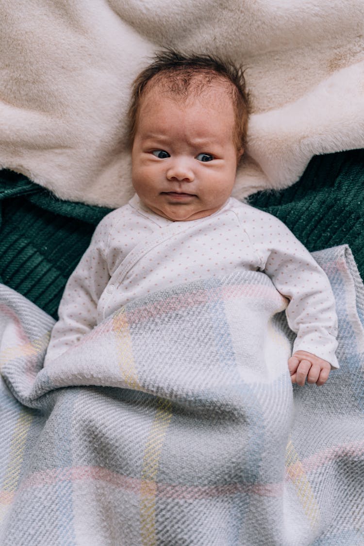 An Infant In Long Sleeves Covered With A Checkered Blanket