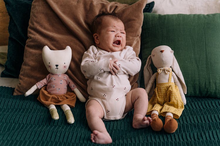 Baby In Pine Onesie Sitting Between Plush Toys