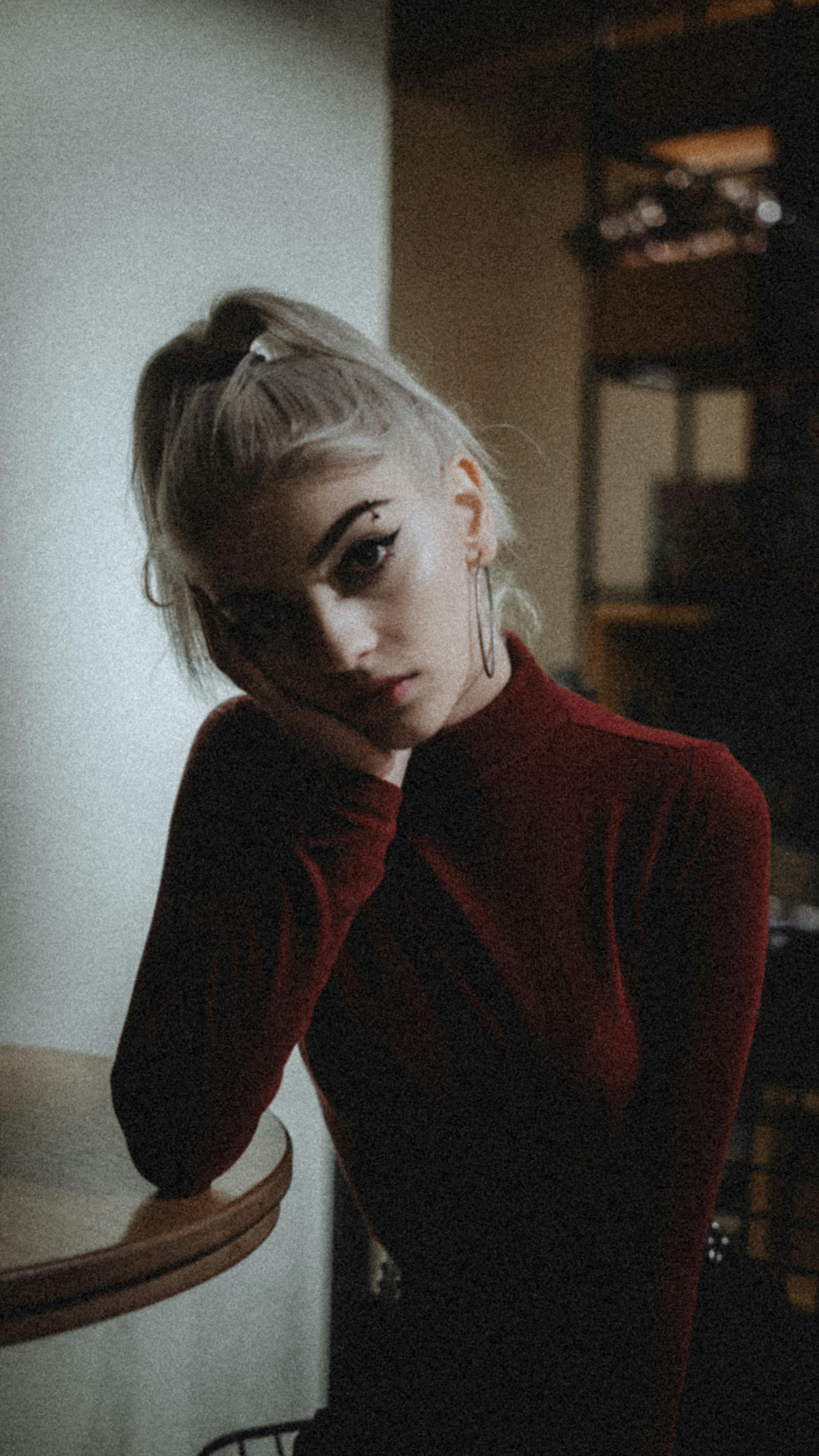 Slim attractive female with ponytail and piercing holding hand on cheek while sitting at table and looking at camera