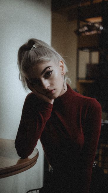 Slim attractive female with ponytail and piercing holding hand on cheek while sitting at table and looking at camera