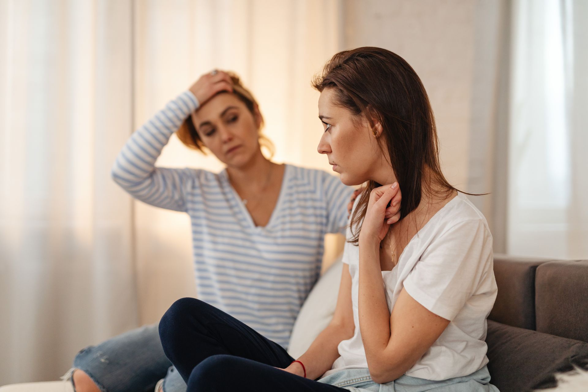 Two women sitting on a sofa showing concern and support for each other indoors.