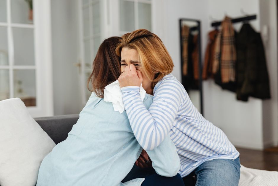 二人の女性が屋内で感情的なサポートを共有しながら抱き合っています。一人の女性は泣いており、もう一人が慰めています。