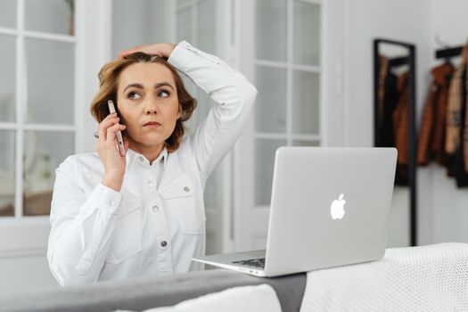Caucasian woman multitasking with smartphone and laptop in modern home office.