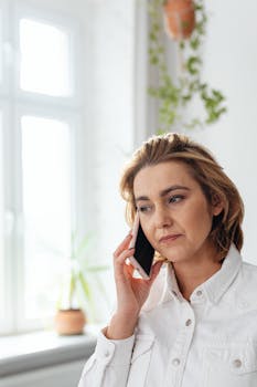 Adult woman in white attire having a phone call indoors with plants in the background.