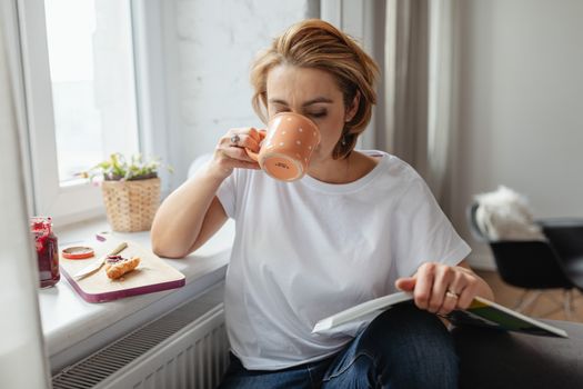 A woman enjoys morning coffee and reading by a bright window.