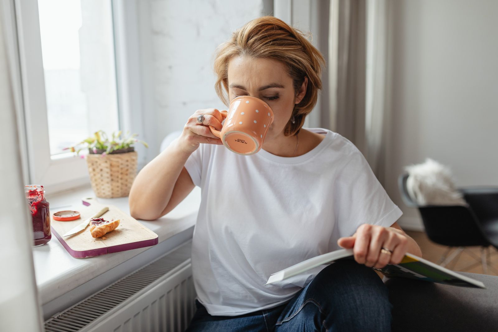 Woman in white T-shirt sips from an orange mug while reading a magazine by a bright window