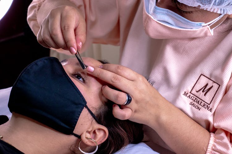 Woman In Pink Uniform Fixing Eyebrow Of A Woman Lying Down And Wearing Face Mask