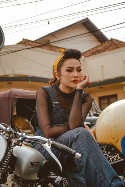 Dreamy millennial Asian female motorcyclist in denim coverall leaning on hand while sitting on motorcycle and looking away in city