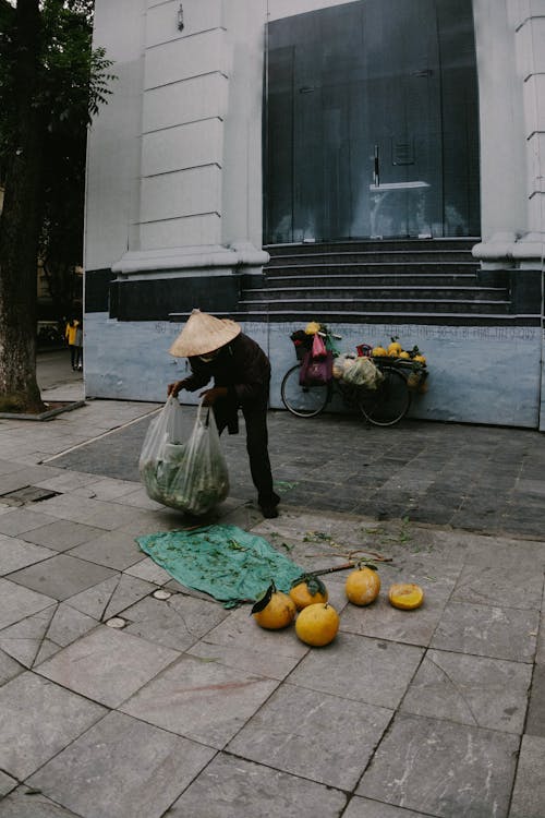 Faceless Asian male fruits seller wearing typical conical hat collecting fruits into plastic bag after working on city street