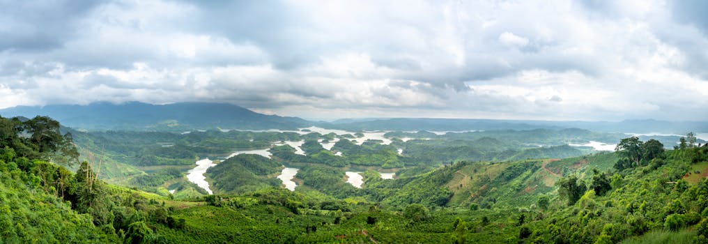 Panorama of curvy river flowing among green terrain in hilly terrain covered with grass against mountain ridge and cloudy sky