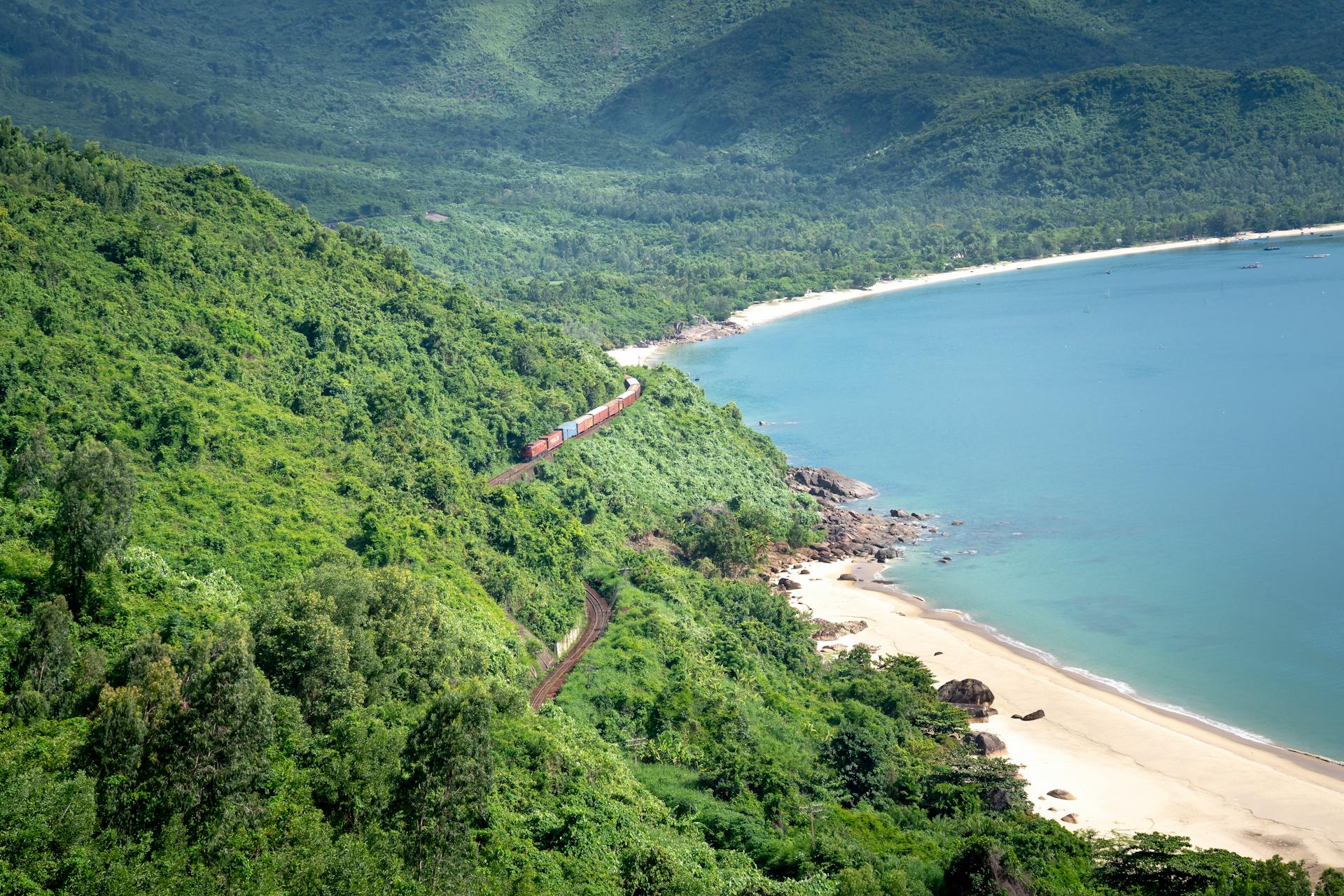 A train travels along a lush, green coastal landscape with a beautiful bay and sandy beach.