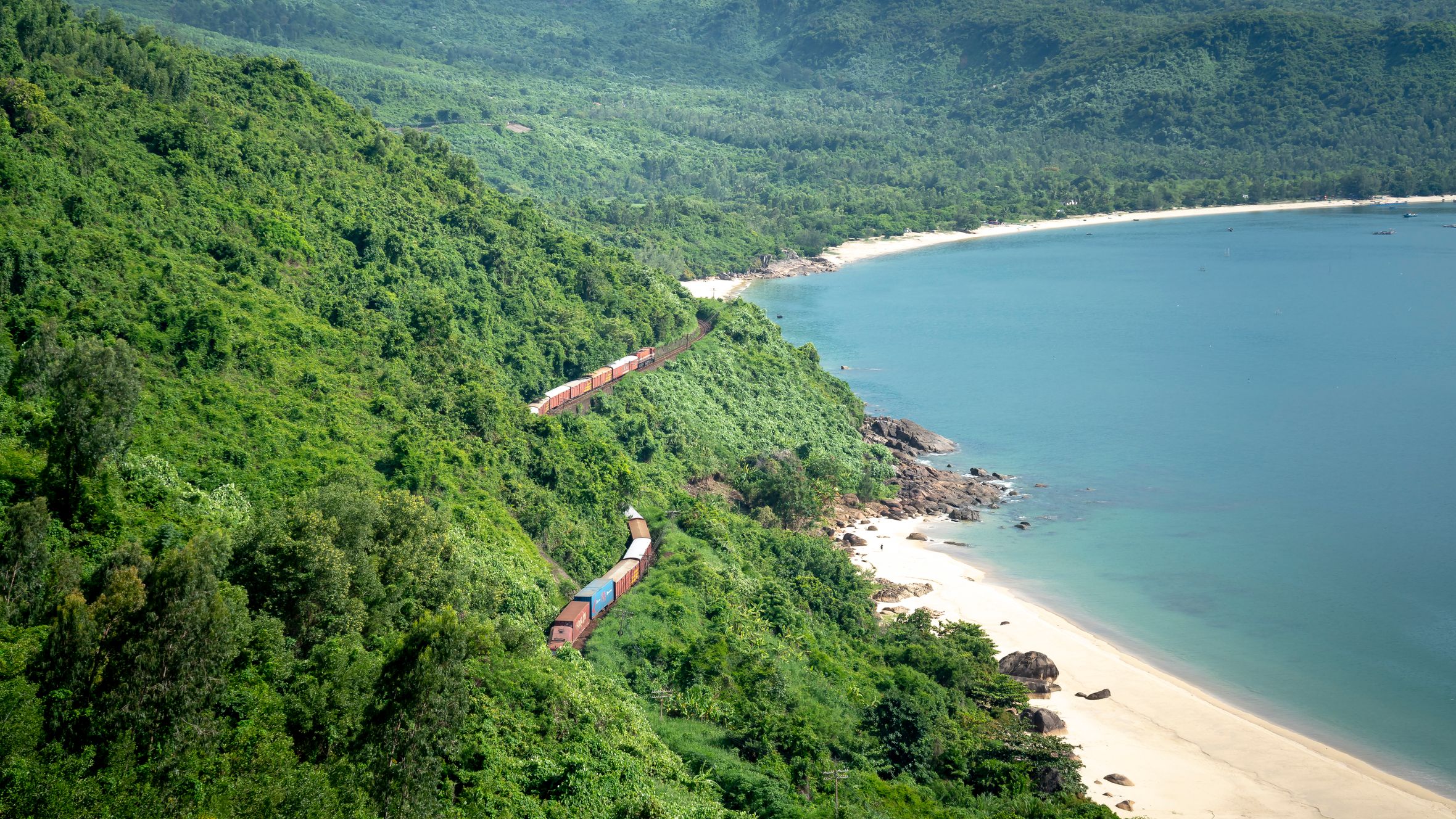 Train going through lush green forest with green trees growing on mountain near sandy shore an calm sea in nature
