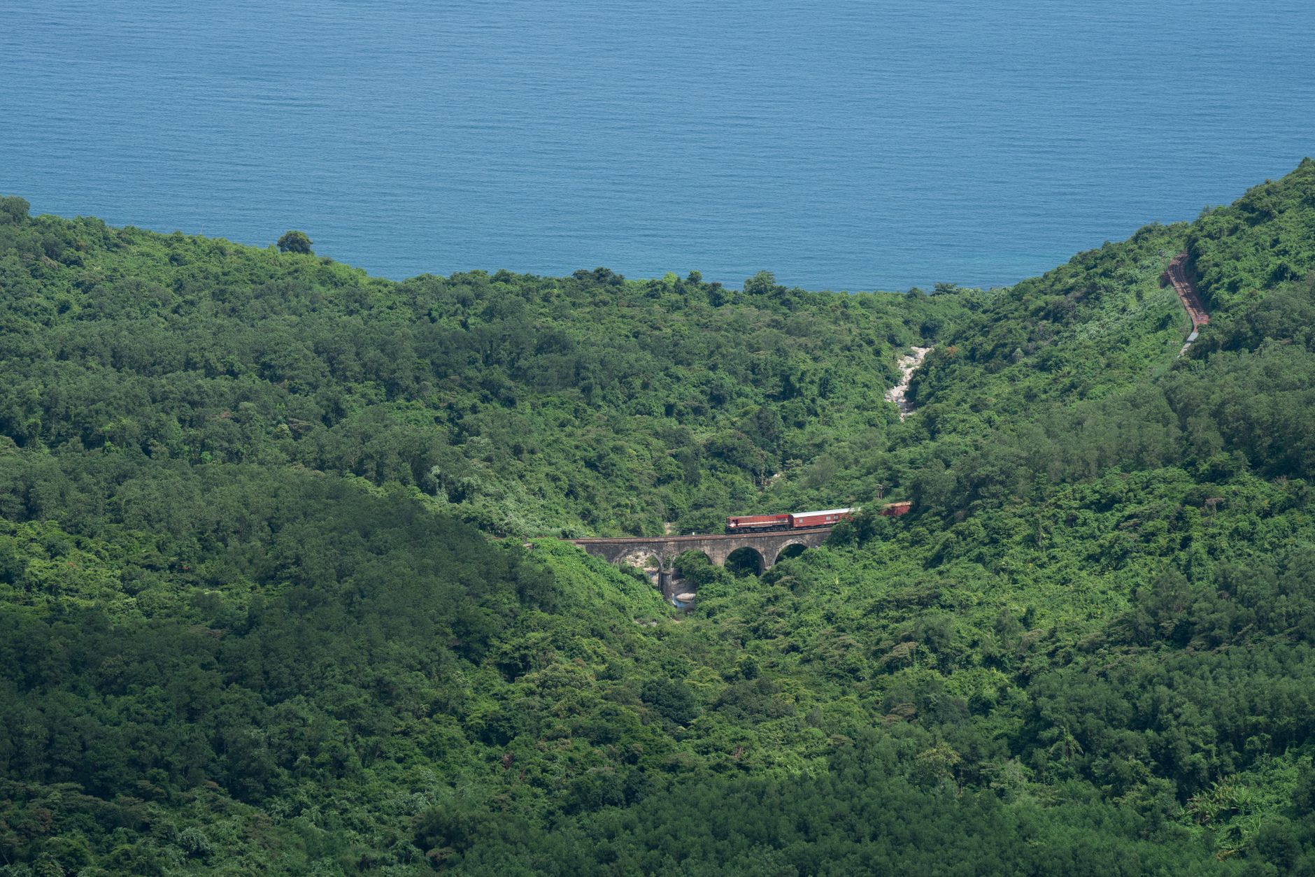 Train riding on old fashioned metal bridge with arched elements through dense forest with green trees against calm endle