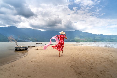Back view full body of unrecognizable female traveler in dress and straw hat strolling on sandy beach near rippling sea against mountain ridge