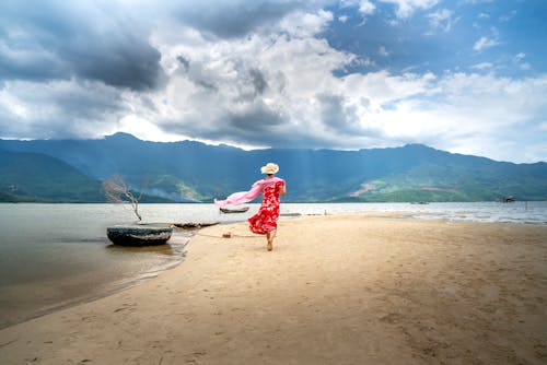 Back view of unrecognizable female traveler with flying textile walking on sandy coast against ocean and ridge under cloudy sky