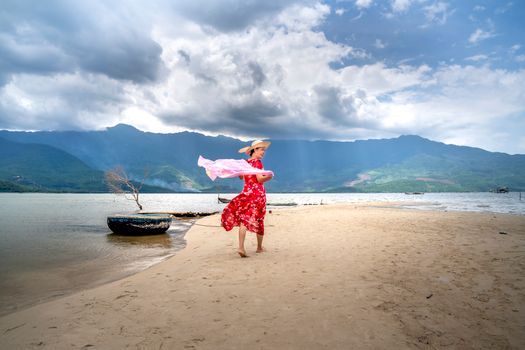 Back view of female tourist with flying textile strolling on ocean coast while looking away against mount under cloudy sky