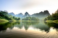 Picturesque landscape of traditional boat floating on rippling lake amidst rocky mountains covered with lush green trees against cloudless sunset sky