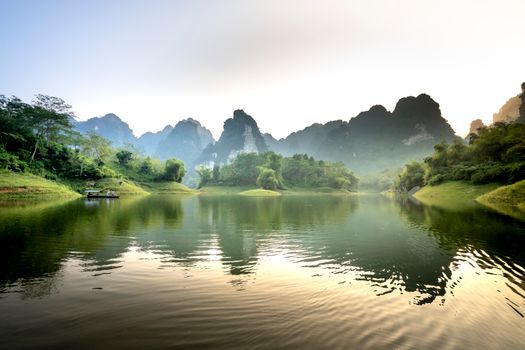 Picturesque landscape of traditional boat floating on rippling lake amidst rocky mountains covered with lush green trees against cloudless sunset sky