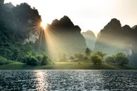 Ripping lake with gassy shore surrounded by massive limestone mountains covered with lush green trees under cloudless sunset sky in Vietnam