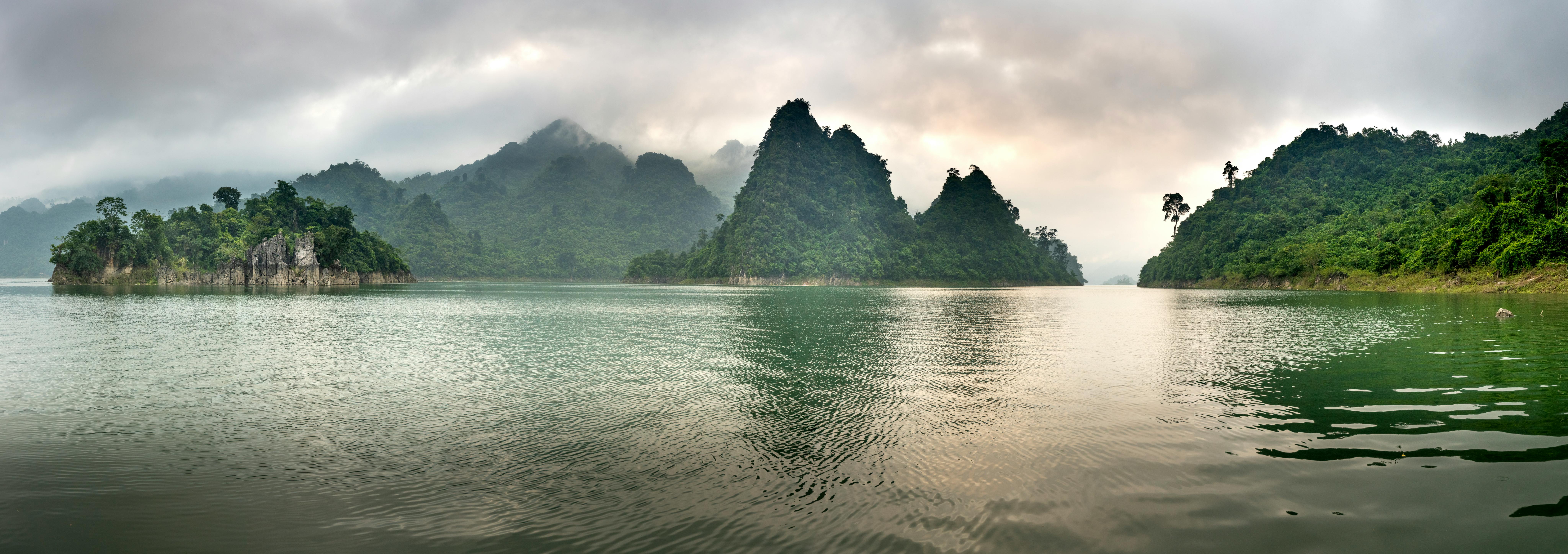 Wide angle scenery of pond surrounded by hills covered with lush tropical forest under gray clouds