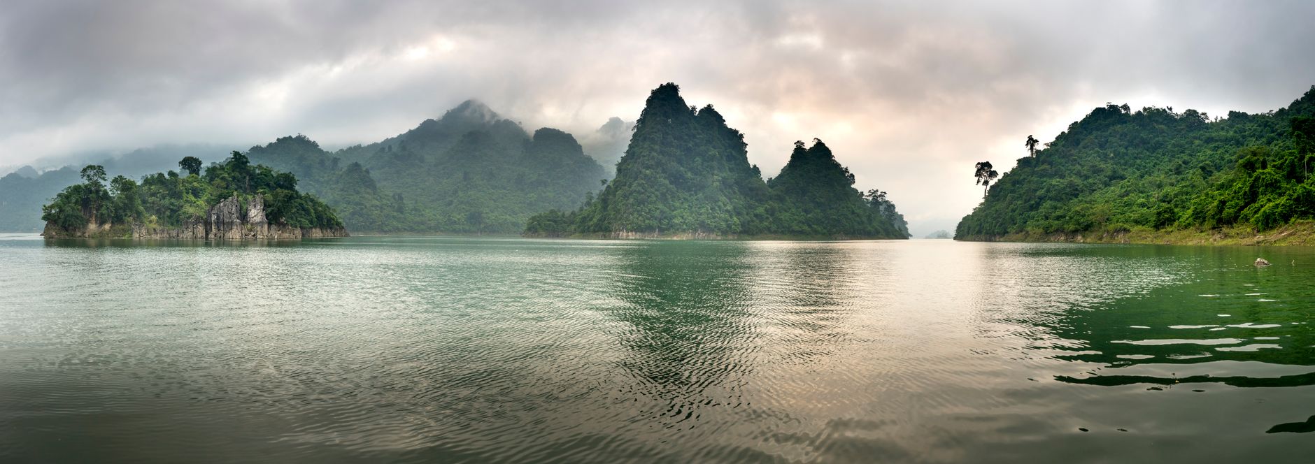 Wide angle scenery of pond surrounded by hills covered with lush tropical forest under gray clouds