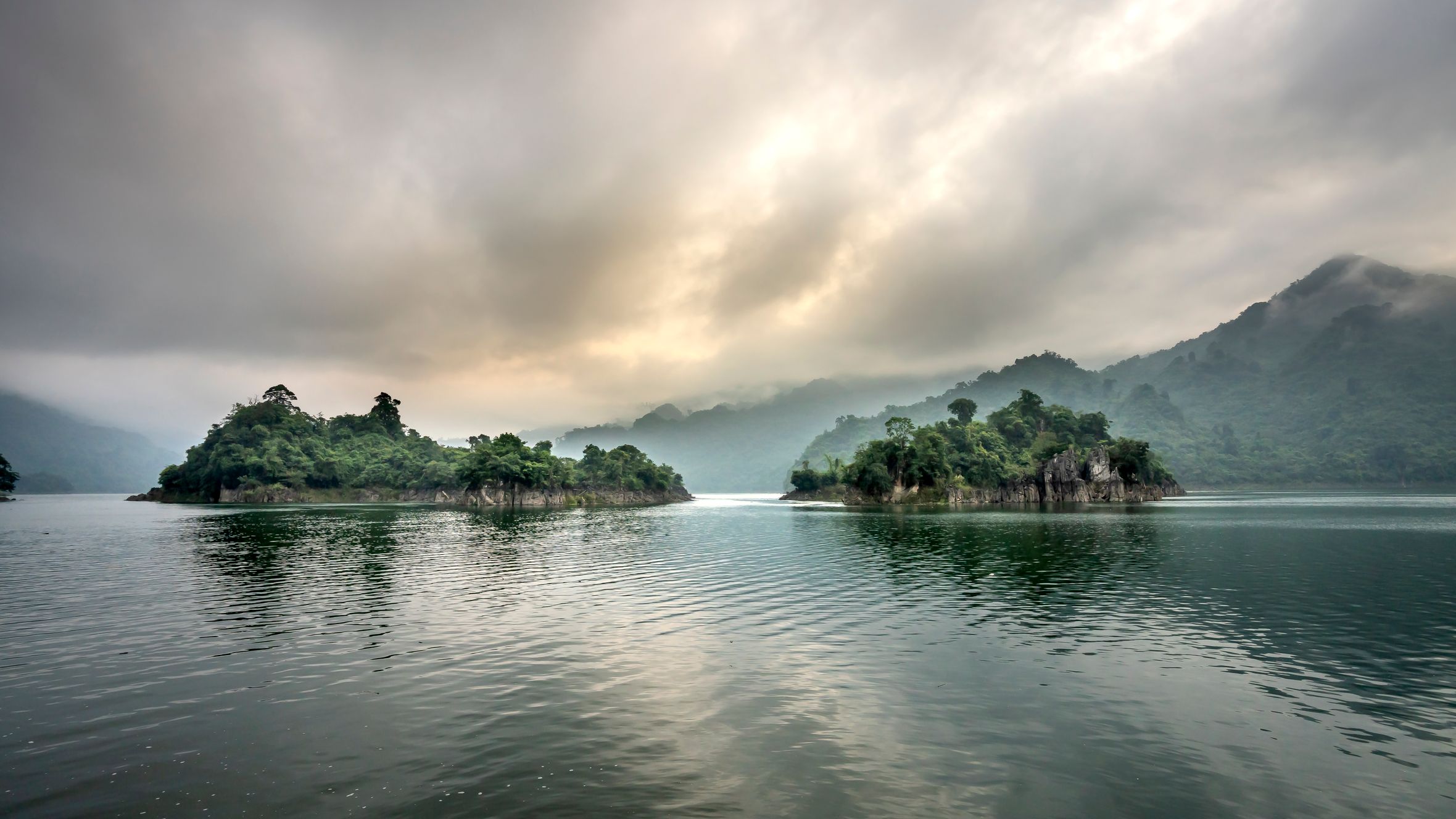 Picturesque scenery of rocky cliffs in rippling water of pond against green hills in mist under clouds