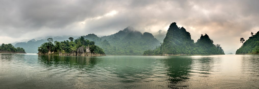 A tranquil panorama of a misty lake surrounded by lush, rocky hills under a cloudy sky.
