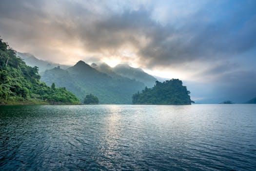 Scenic view of foggy mountains with lush trees against wavy sea under shiny cloudy sky in evening