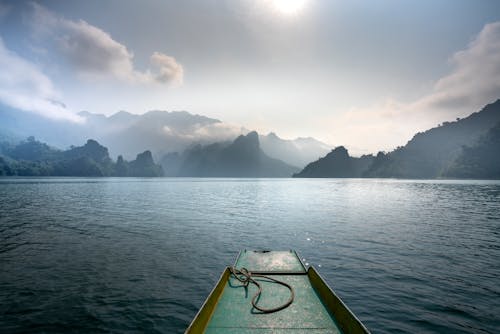 Scenic view of boat on rippled ocean against mounts in mist under cloudy sky with shiny sun