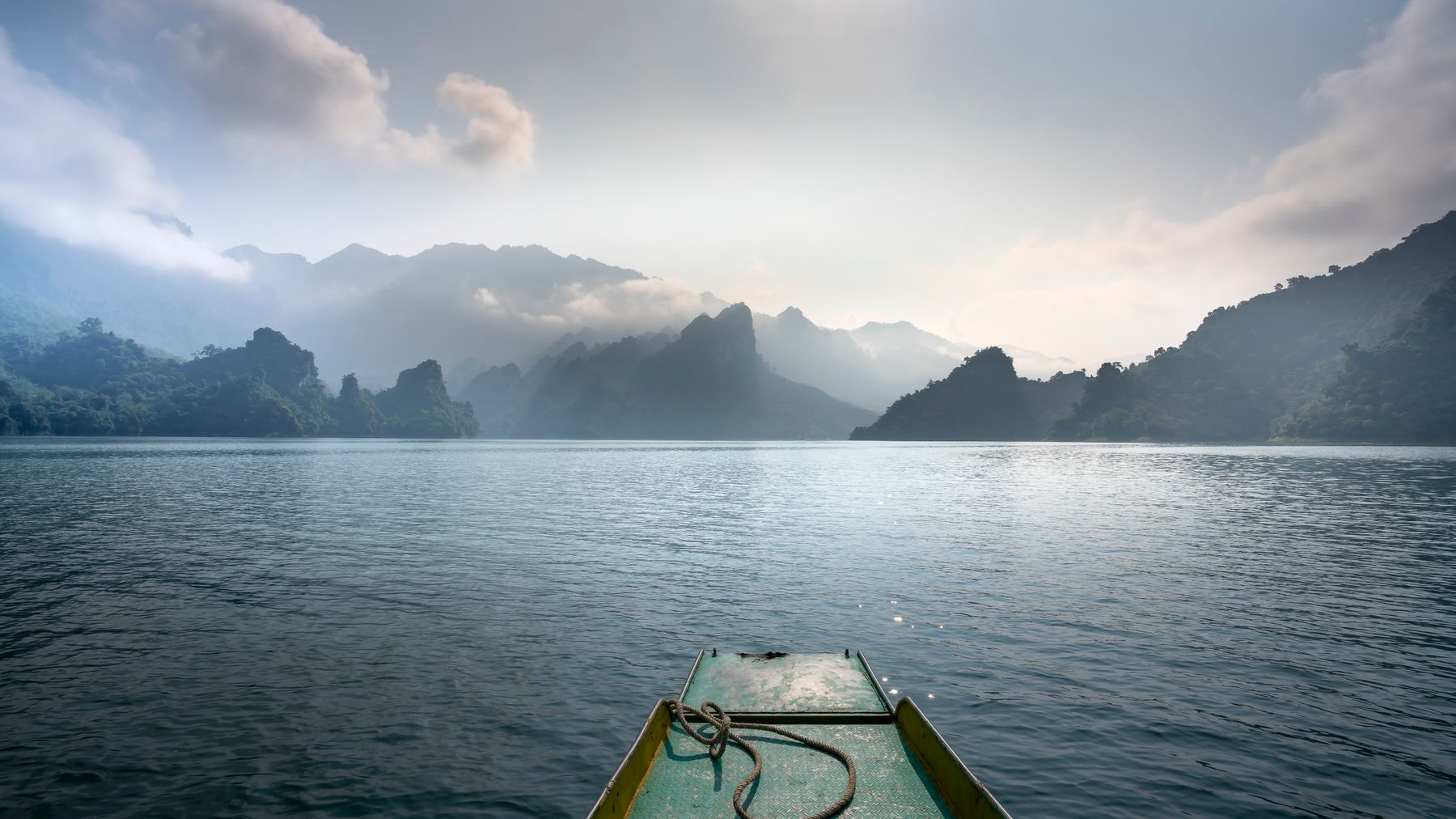 Scenic view of boat on rippled ocean against mounts in mist under cloudy sky with shiny sun