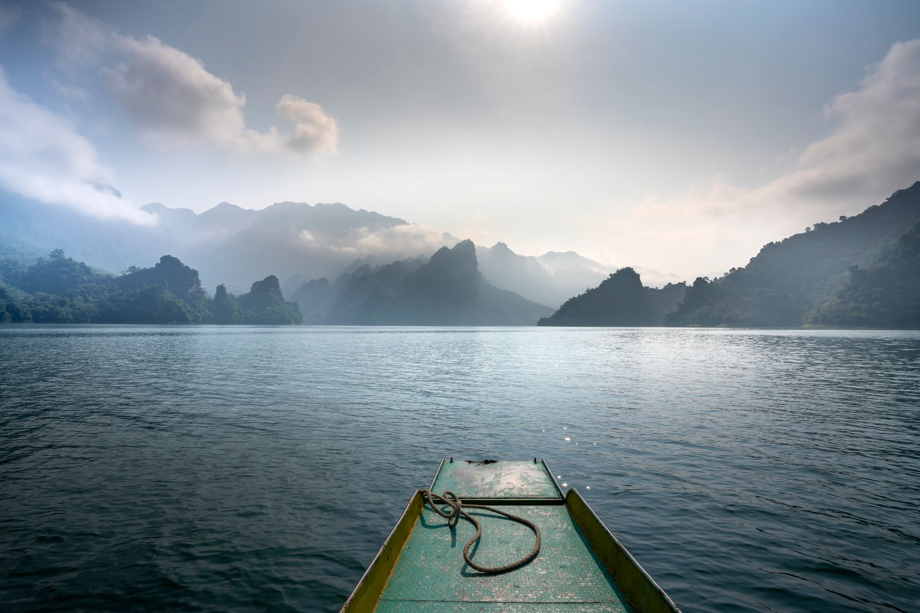 Scenic view of boat on rippled ocean against mounts in mist under cloudy sky with shiny sun