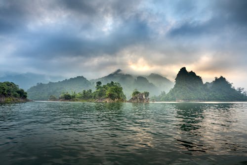 Free Picturesque view of mounts with lush trees against rippled sea under fluffy clouds at sunset in misty weather Stock Photo