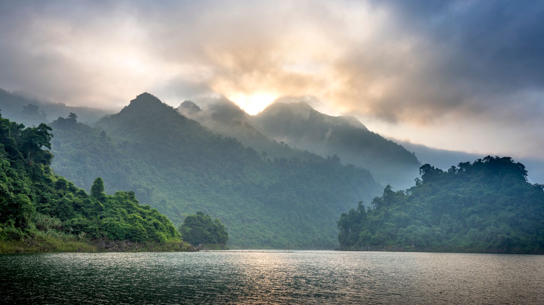 Spectacular view of ridges with lush green trees against ocean under cloudy sky at sunset in foggy weather