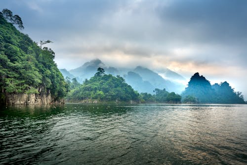 Scenery view of majestic mountains with lush green trees against rippled ocean under fluffy clouds at sunset in misty weather