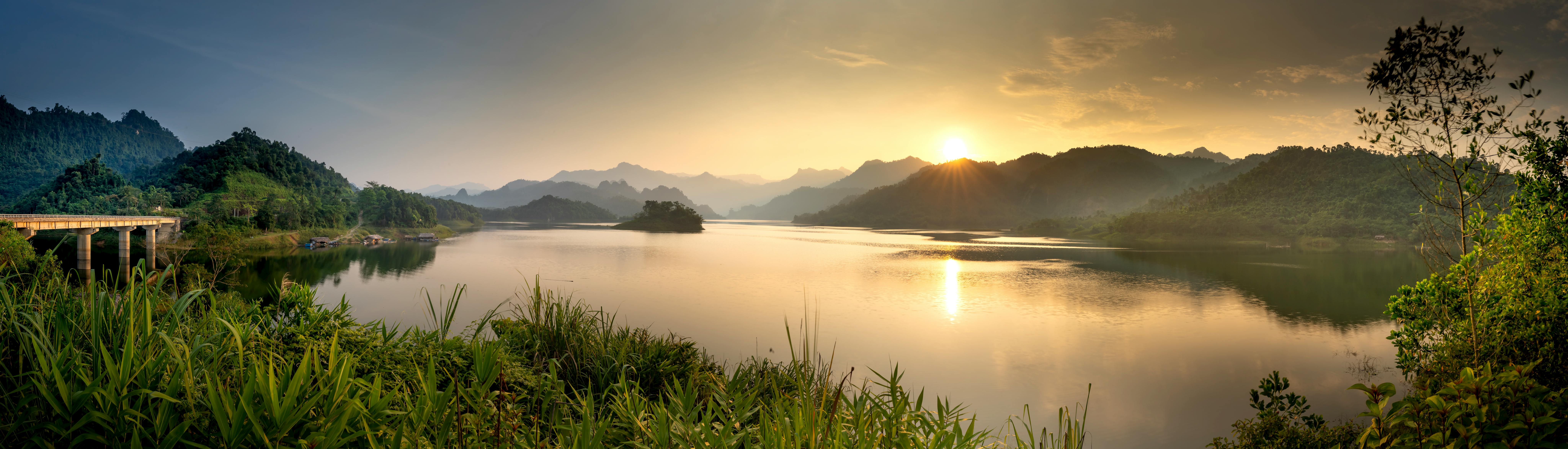 Panoramic view of lake against high mountains and green plants illuminated by sun in twilight