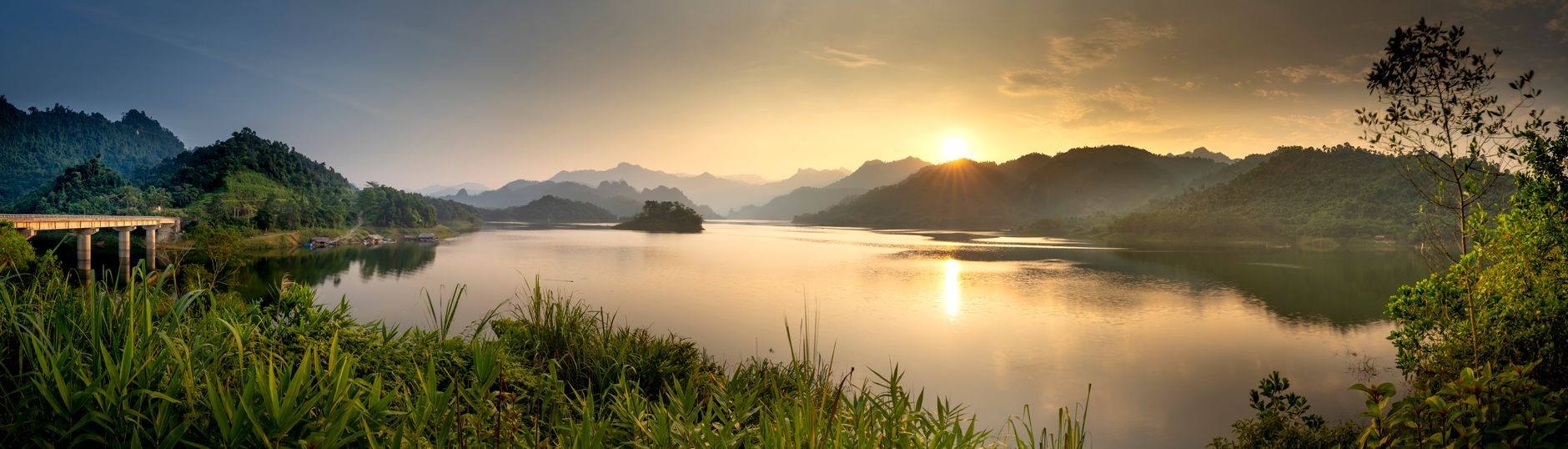 Panoramic view of lake against high mountains and green plants illuminated by sun in twilight