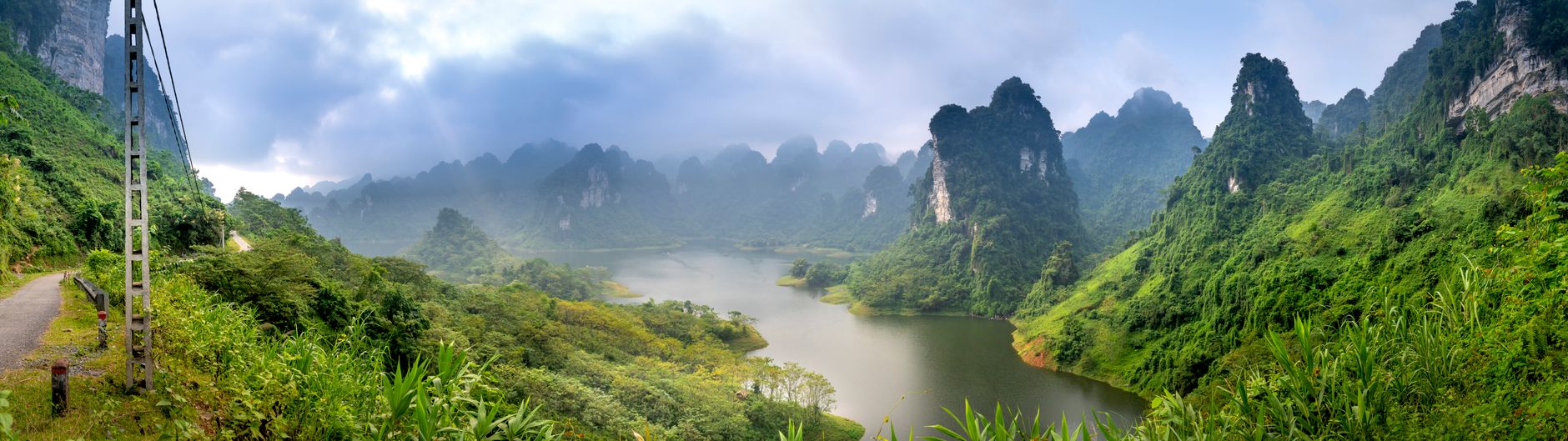 Wide angle of lake between high mounts with lush green trees and roadway under cloudy sky in misty weather